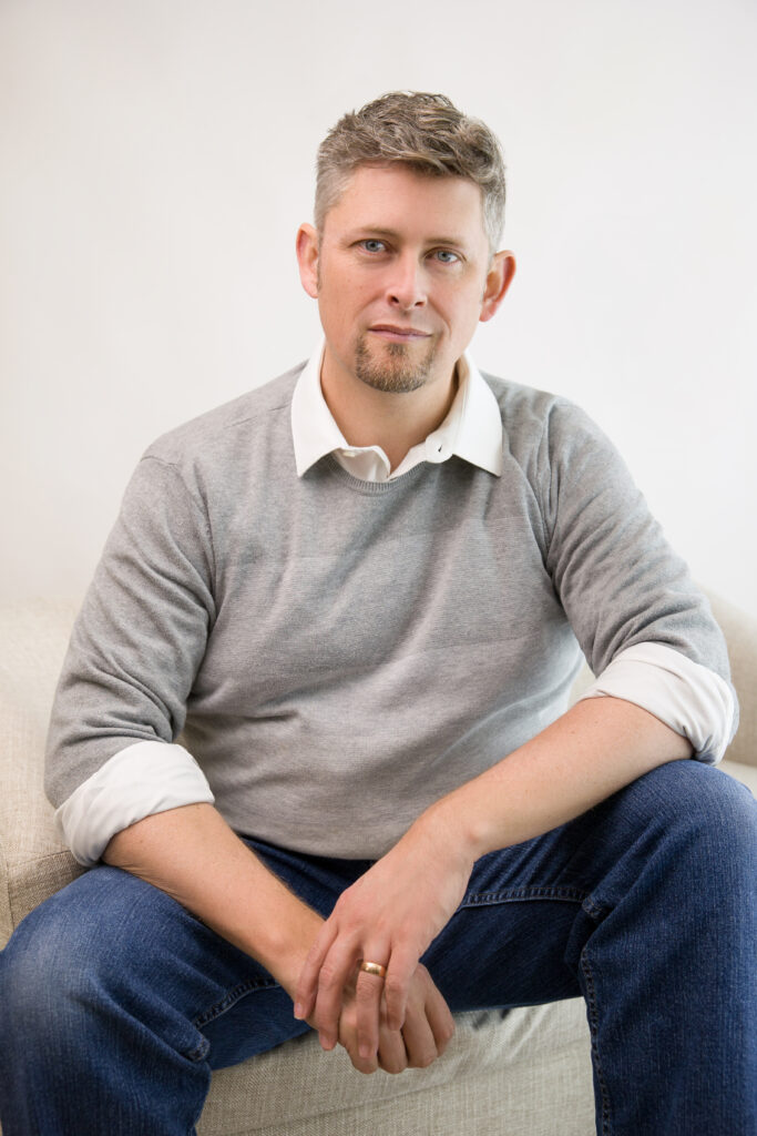 man on couch posing for headshot photography in grey striped sweater, white collared shirt, blue jeans 