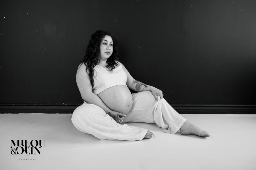 Black and white maternity portrait of a pregnant woman seated on the floor, wearing light linen pants and a sleeveless top against a dark studio backdrop.