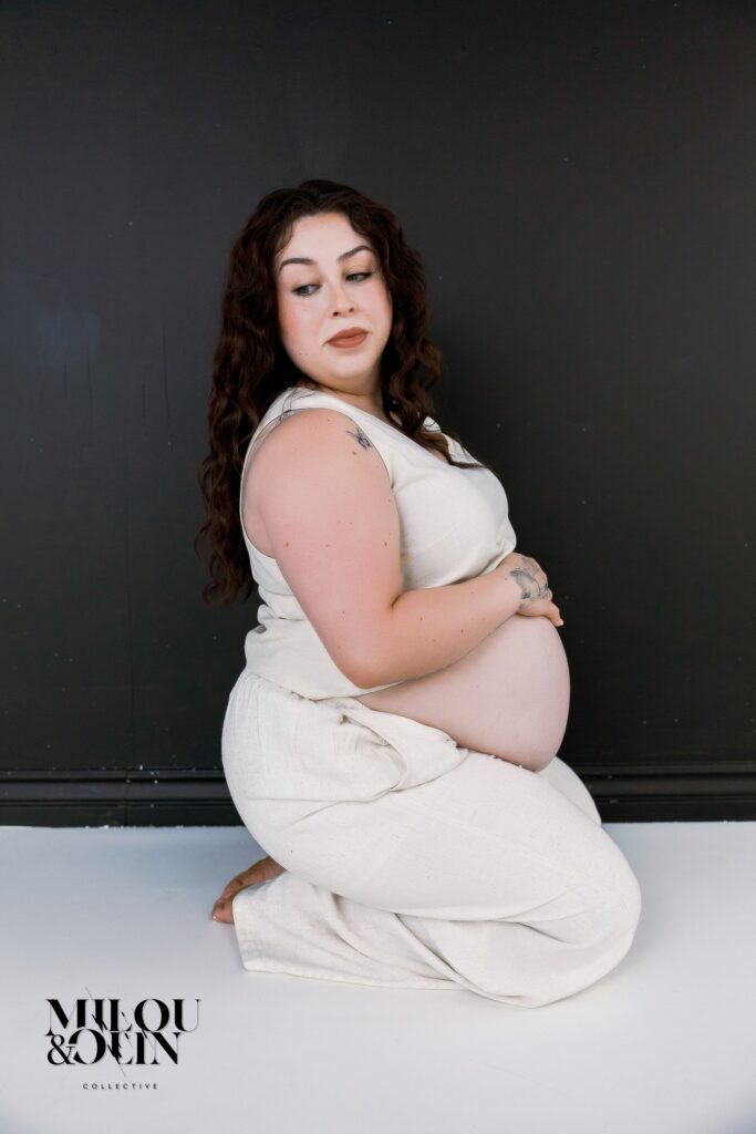 Maternity portrait of a pregnant woman seated on the floor in soft neutral clothing, resting her hands on her belly against a dark studio wall.