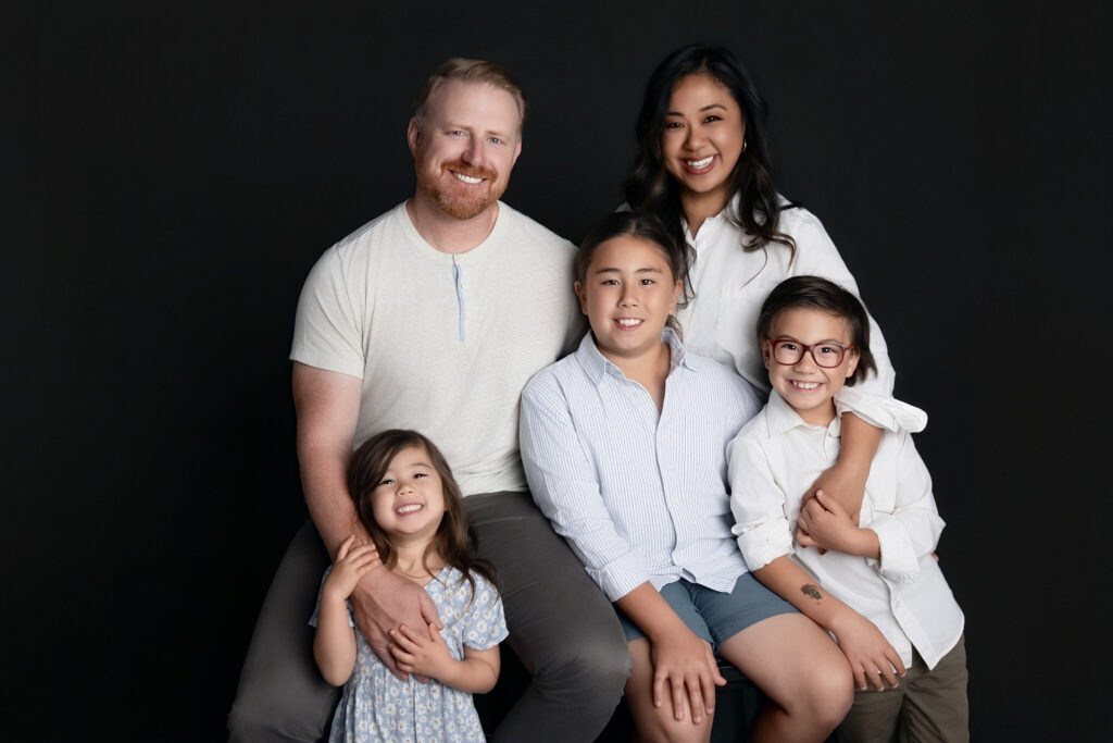 Family of five posing for a studio portrait against a dark backdrop, featuring coordinated neutral outfits, joyful expressions, and close family connection.