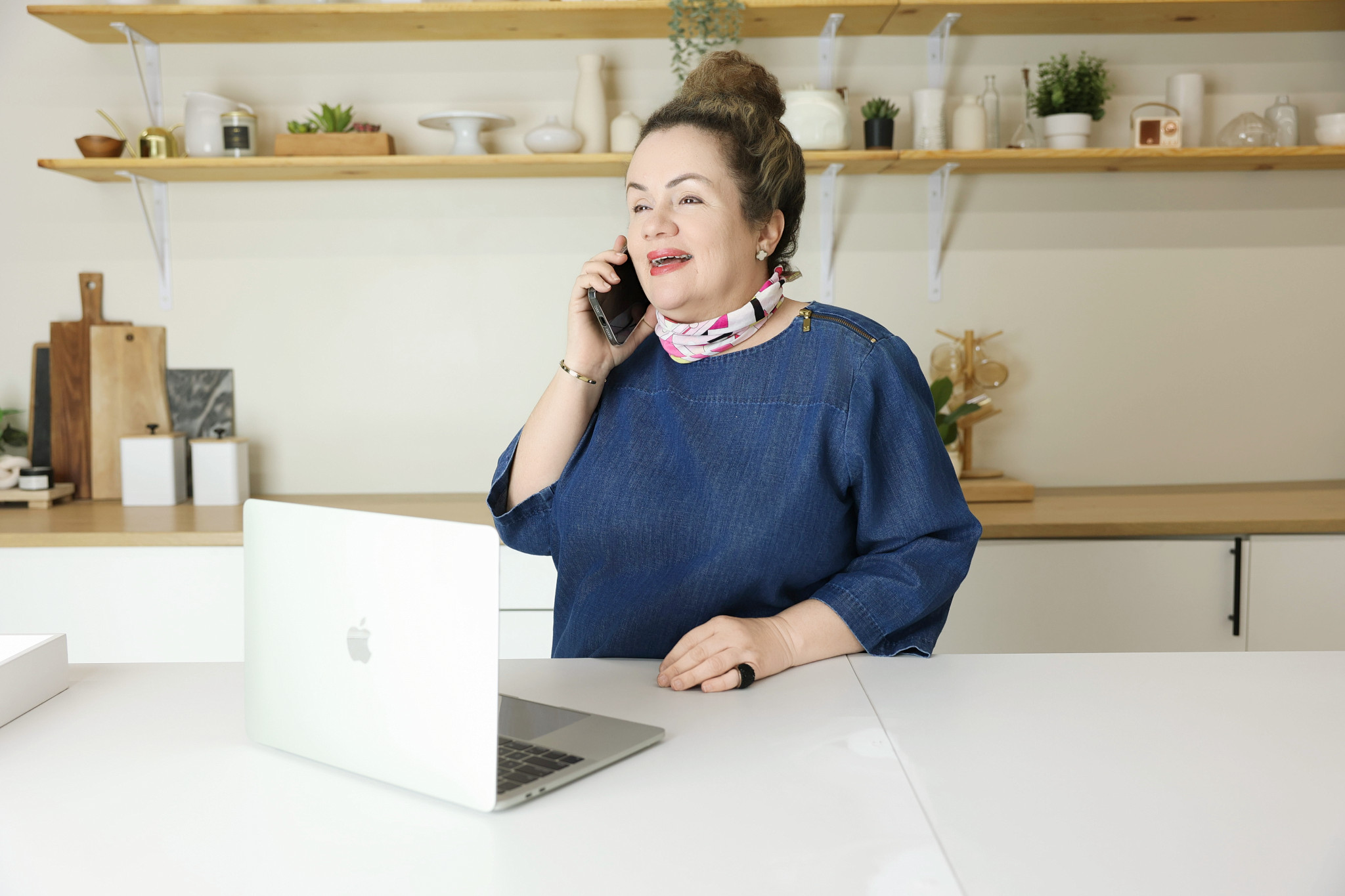 Professional woman standing in a modern kitchen workspace while talking on the phone beside an open laptop, surrounded by clean shelves, neutral decor, and natural light.