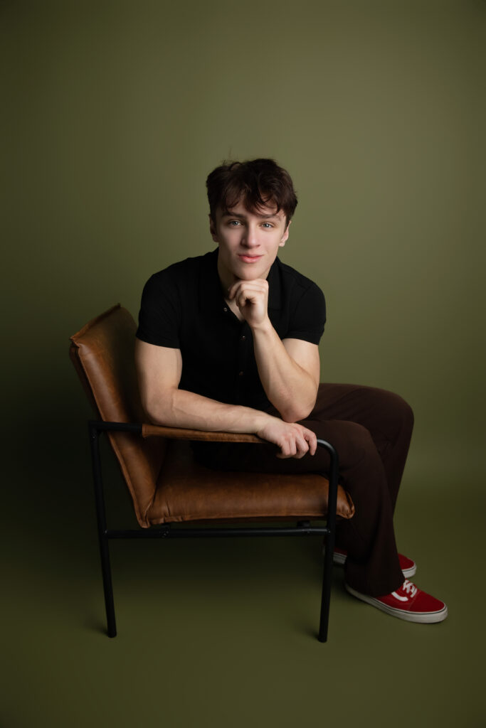 Modern Sacramento senior portrait of a teenage boy posing in a leather chair with a relaxed editorial style and moody olive backdrop by Milou & Olin Collective.