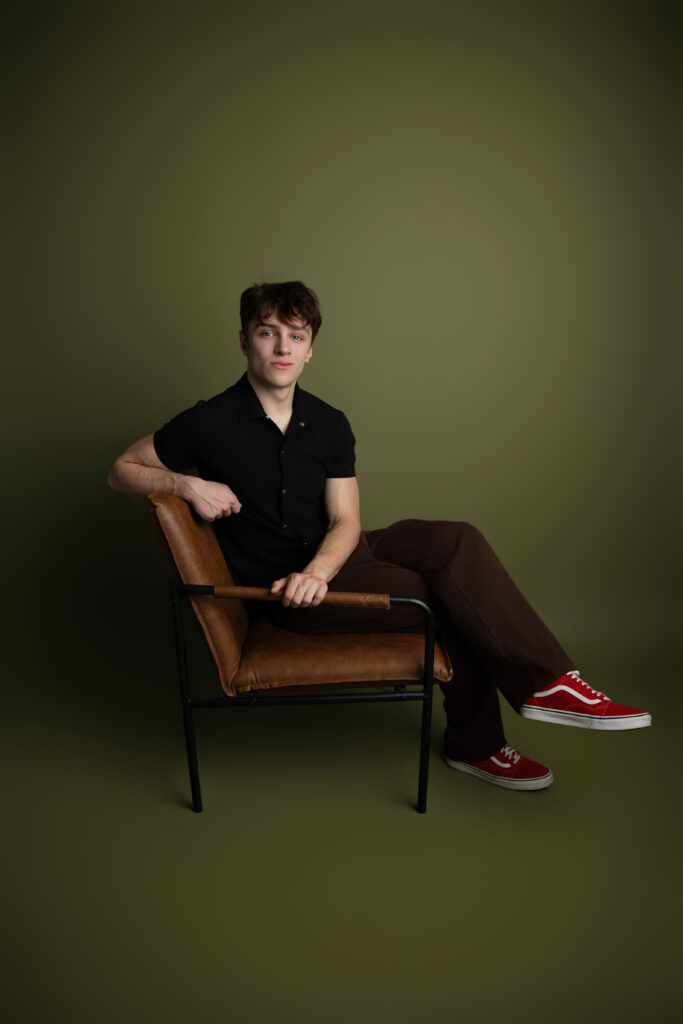 Editorial senior portrait photography featuring a teenage boy seated in a modern leather chair with cinematic lighting and bold red sneakers in a Sacramento studio.