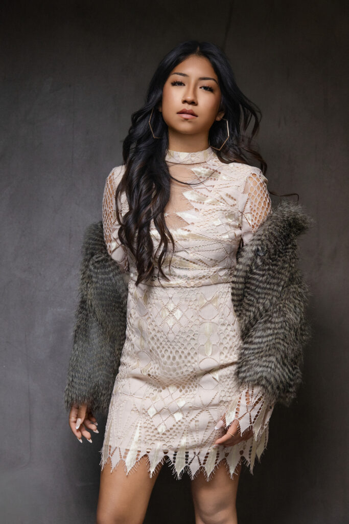 Fashion-forward senior portrait of a teenage girl wearing a textured ivory dress and faux fur jacket, photographed with editorial lighting in a Sacramento studio.