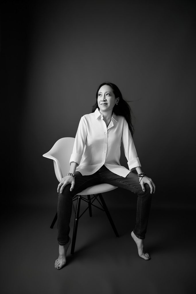 Elegant black-and-white portrait of a woman seated barefoot on a modern white chair, wearing a crisp white shirt and jeans during a breast cancer portrait session Sacramento.