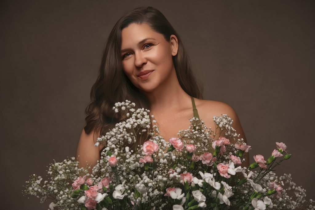 Luxury editorial portrait of a woman surrounded by delicate pink carnations and white flowers against a muted brown backdrop during a breast cancer portrait session Sacramento.