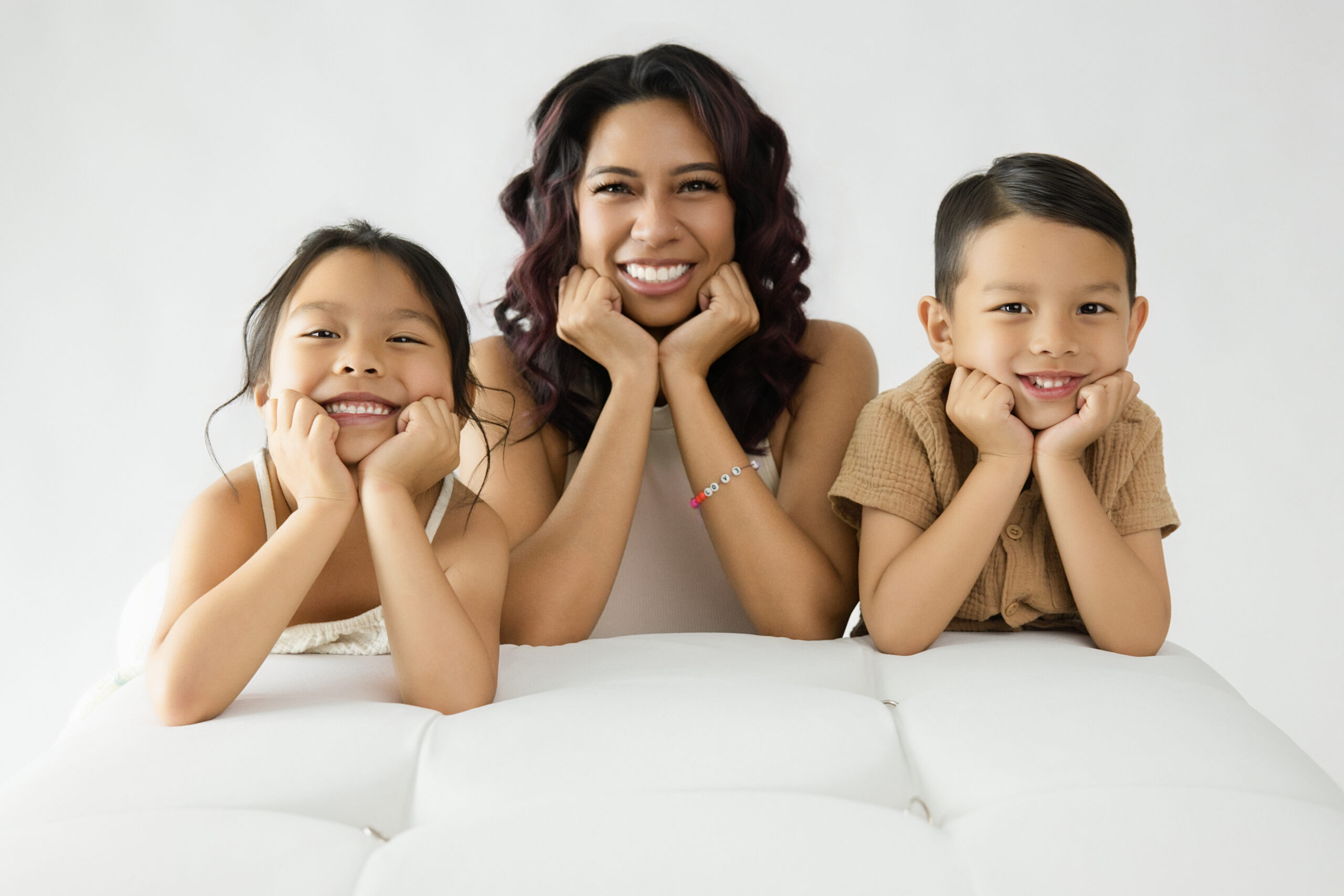 Luxury Sacramento family portrait photography featuring a smiling mother with her daughter and son, all resting their chins on their hands while posing together on a white tufted sofa against a bright, minimalist studio backdrop.